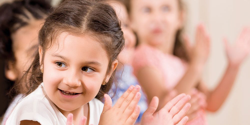 Children clap in early learning setting. Camera focuses on girl with brown hair, other kids in the background are blurred